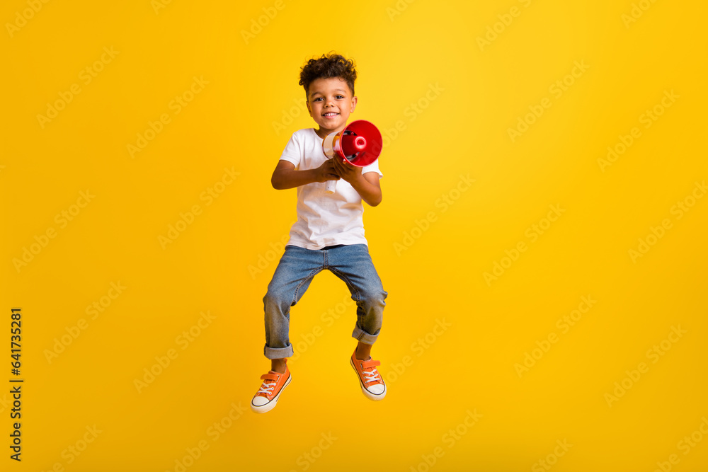 Full length photo of cute pretty small boy wear white t-shirt jumping high screaming bullhorn isolated yellow color background Full length photo of cute pretty small boy wear white t-shirt jumping high screaming bullhorn isolated yellow color background