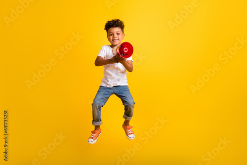 Full length photo of cute pretty small boy wear white t-shirt jumping high screaming bullhorn isolated yellow color background