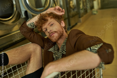 Obraz na płótnie young stylish redhead man in jacket looking at camera while sitting in cart in c