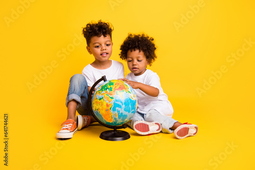 Full body portrait of cute intelligent curious brothers sit floor look planet earth globe isolated on yellow color background