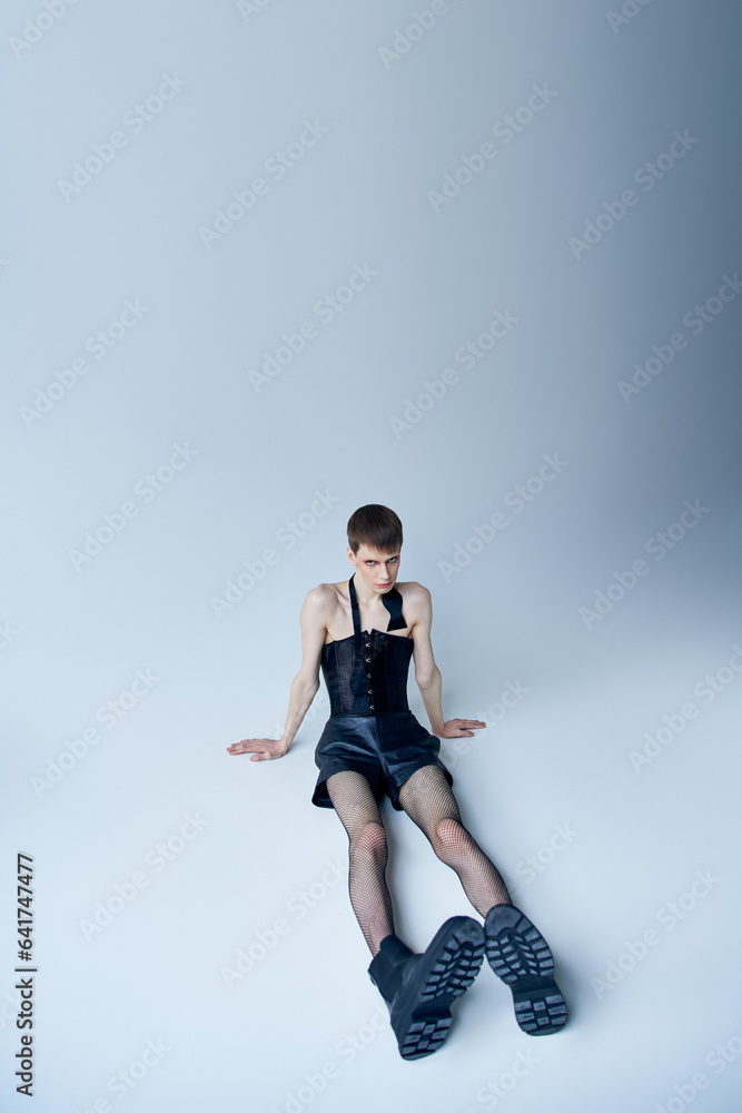 queer person in black corset and fishnet tights sitting on grey, androgynous model, lgbt fashion