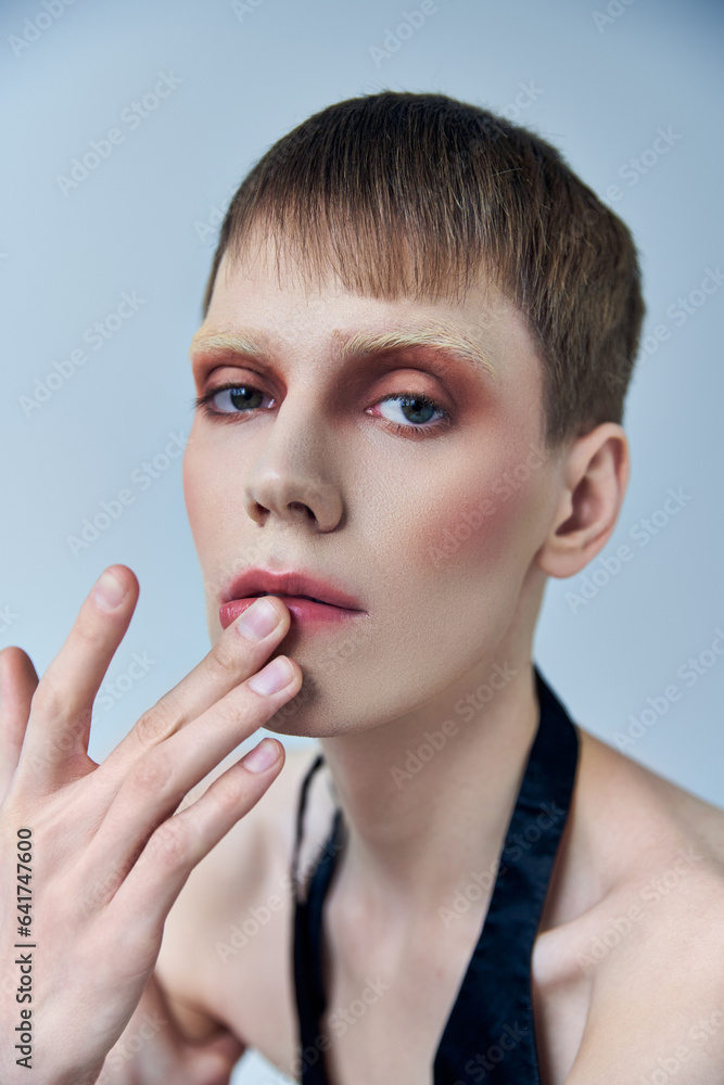 queer person with makeup on grey backdrop, androgynous person, touching lip, look at camera queer person with makeup on grey backdrop, androgynous person, touching lip, look at camera