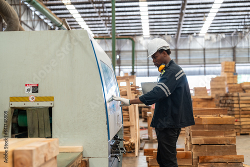 Black man worker operating CNC machine for cutting wood in factory. Male engineer in uniform with helmet safety working produce wooden pallet at manufacturing plant.