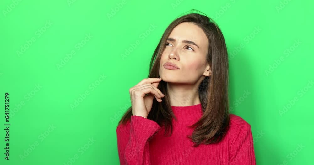 Pensive puzzled brunette young girl wearing pink sweater posing isolated on green background in studio. Looking aside up put hand prop up on chin, lips. Thoughtful woman, grimacing thinking girl