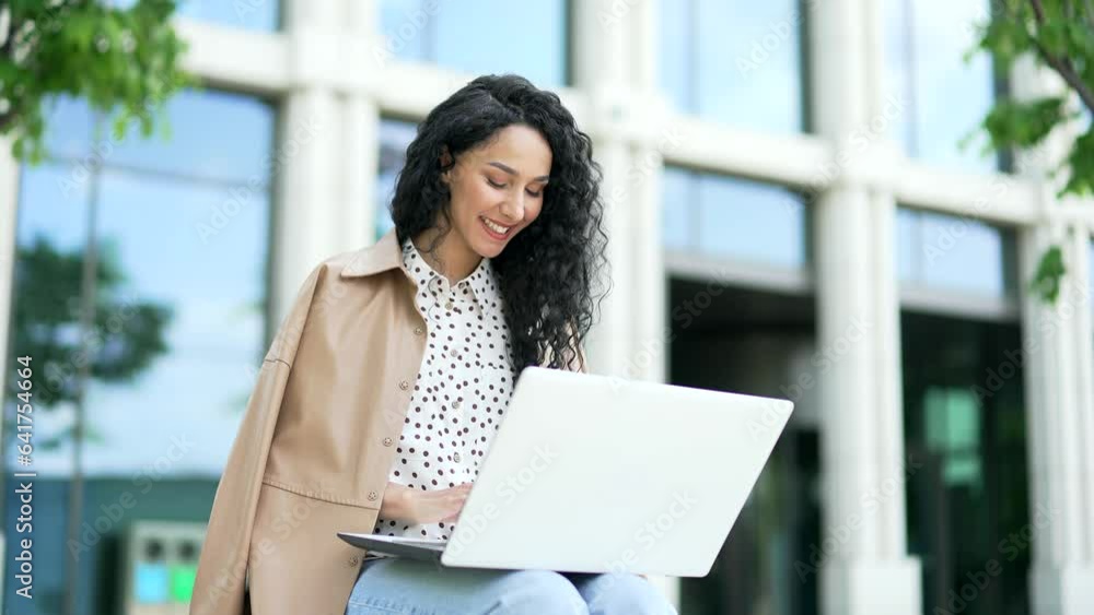 Young female entrepreneur works typing on a laptop while sitting on a bench near an office building. Freelancer chatting online, typing a message, checking email, corresponding remotely with a client Young female entrepreneur works typing on a laptop while sitting on a bench near an office building. Freelancer chatting online, typing a message, checking email, corresponding remotely with a client
