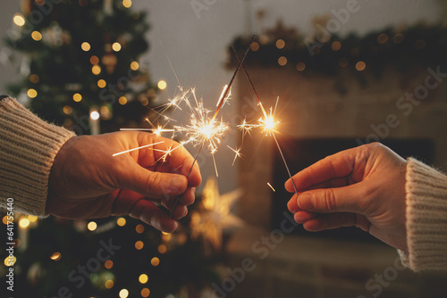 Hands holding burning fireworks against modern fireplace and christmas tree with golden lights Fototapeta