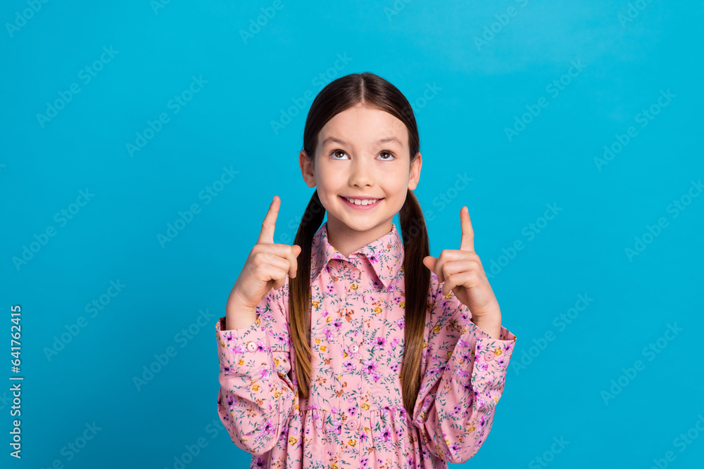 Photo portrait of pretty schoolgirl look point up empty space dressed stylish pink flower print outfit isolated on blue color background Photo portrait of pretty schoolgirl look point up empty space dressed stylish pink flower print outfit isolated on blue color background