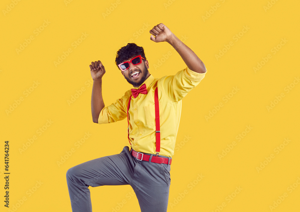 Happy funny joyful young South Asian Indian man wearing yellow shirt, red bow tie, sunglasses and grey trousers with suspenders celebrating, having fun and dancing on yellow background. Party concept Happy funny joyful young South Asian Indian man wearing yellow shirt, red bow tie, sunglasses and grey trousers with suspenders celebrating, having fun and dancing on yellow background. Party concept