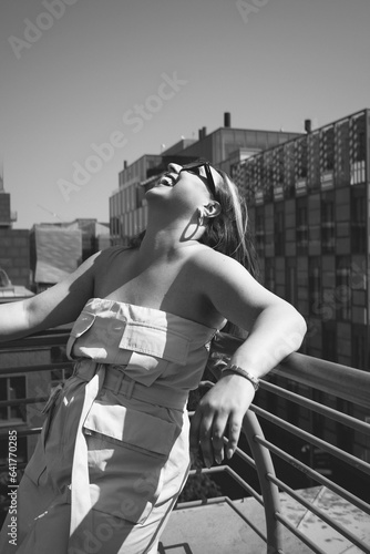Joyful Heights: B&W Rooftop Portrait of Laughing Curvy Model with Sunglasses & Cityscape