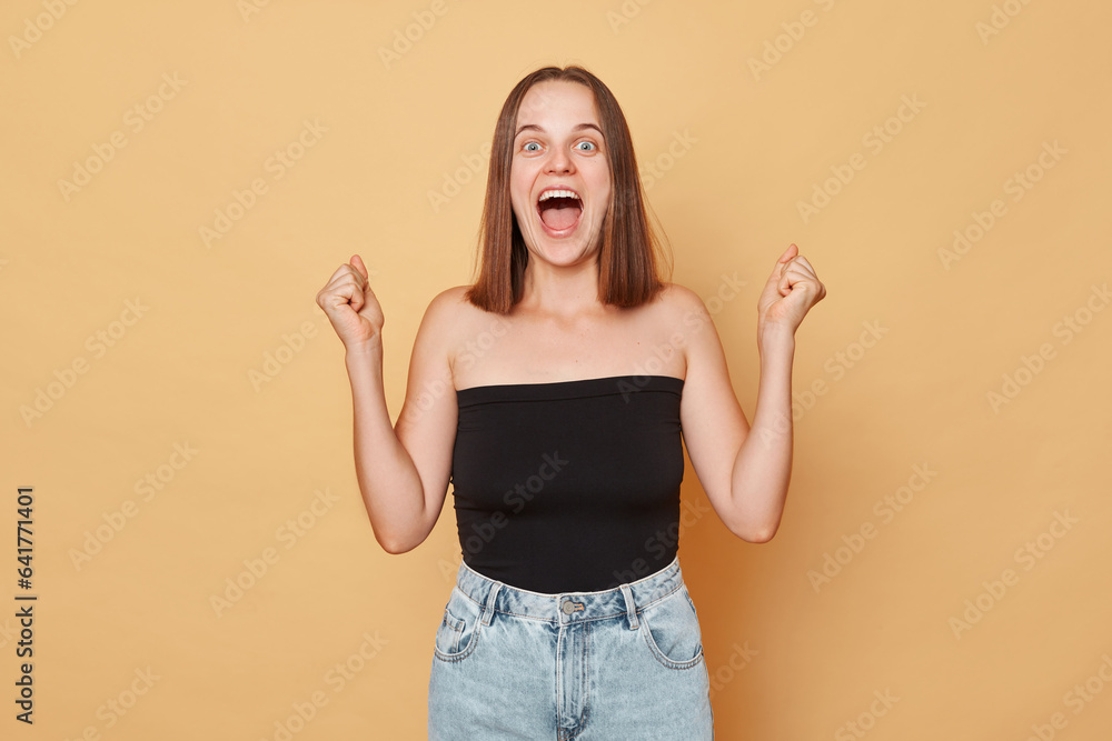 Overjoyed surprised brown haired young woman wearing black top and jeans standing isolated over beige background celebrfating her success clenched fists Overjoyed surprised brown haired young woman wearing black top and jeans standing isolated over beige background celebrfating her success clenched fists