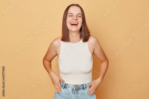 Laughing positive brown haired young woman wearing casual clothing standing isolated over beige background lookign at camera hearing funny joke expressing positive emotions © sementsova321