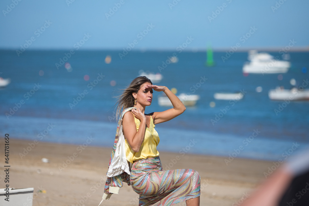 Beautiful young blonde woman looks at the horizon on the seashore, shielding herself from the sun with her hand. In the background on the horizon the blue sea and the boats at sea. Holiday concept.