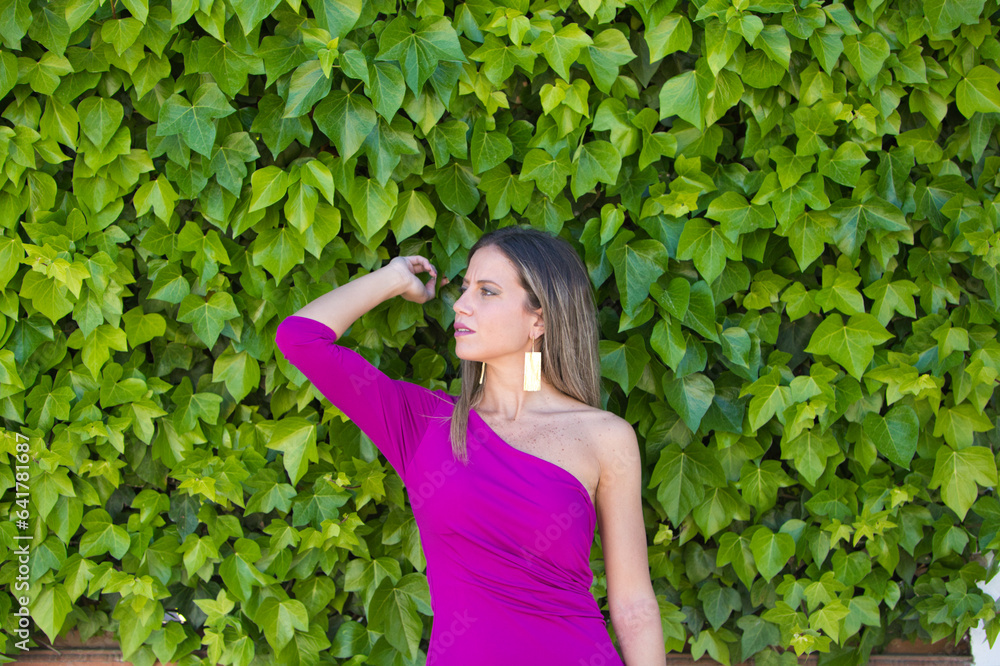 Pretty young blonde woman in purple dress posing for photos against background of a wall of ivy. Woman makes different expressions, serious, happy. Pretty young blonde woman in purple dress posing for photos against background of a wall of ivy. Woman makes different expressions, serious, happy.