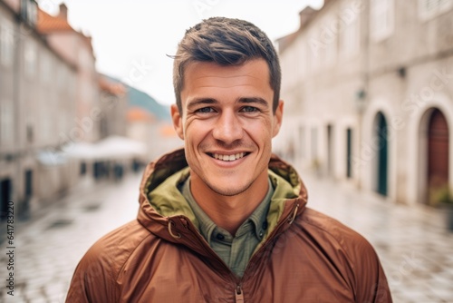 Headshot portrait photography of a grinning boy in his 30s wearing a lightweight packable anorak at the dubrovnik old town in dubrovnik croatia. With generative AI technology
