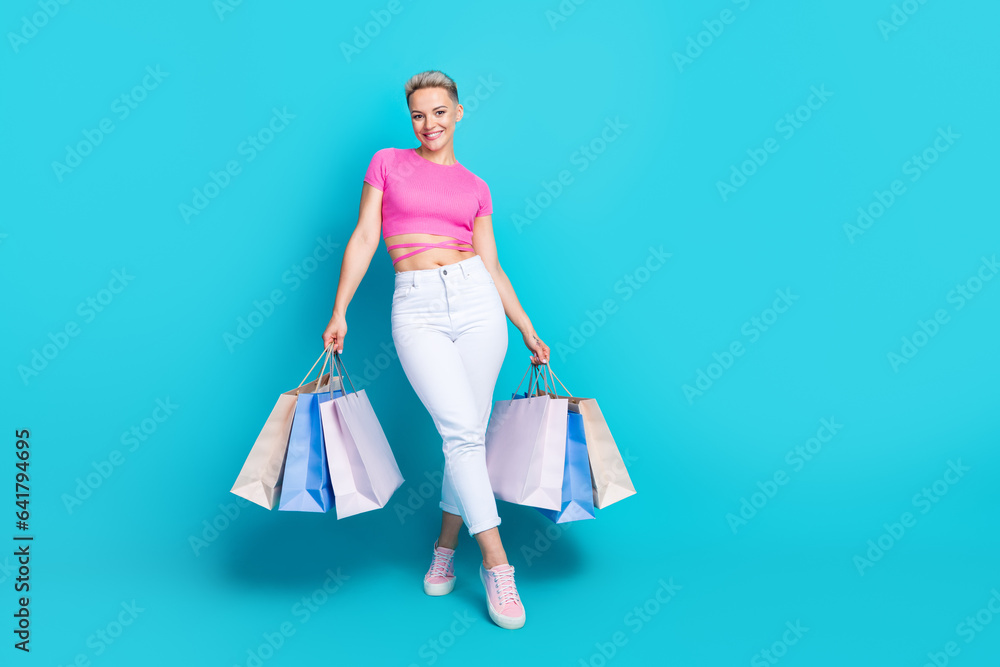 Full size photo of cute lovely good mood woman wear pink top white trousers holding shopping bags isolated on blue color background Full size photo of cute lovely good mood woman wear pink top white trousers holding shopping bags isolated on blue color background