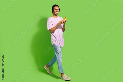 Full length photo of funny charming man dressed purple t-shirt texting modern device walking empty space isolated green color background © deagreez