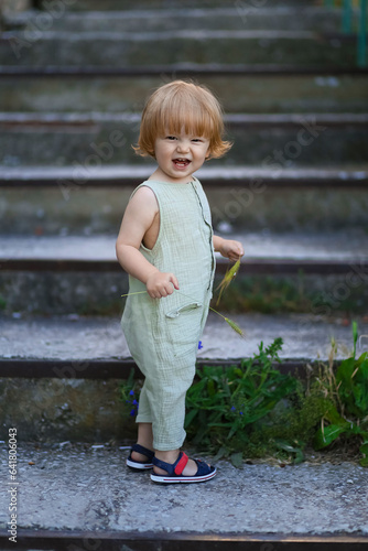 little cute baby boy blond in linen overalls stands on the stairs in the park on a summer day, children's natural cotton clothes, emotional toddler, country lifestyle, farm
