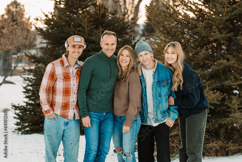 family with adult children standing together in winter wear in the snow in front of a bunch of pine trees