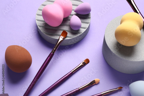 Decorative podiums with makeup brushes and sponges on lilac background