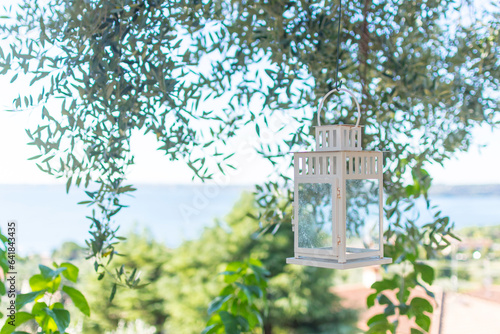 A white lantern hangs in a lounge from an olive tree. In the background Lake Garda on a sunny summer day. In the evening the lantern spreads cozy atmosphere.