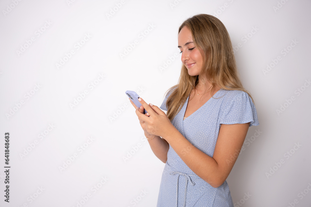 Happy woman texting on a smartphone looking at phone on a white wall Happy woman texting on a smartphone looking at phone on a white wall