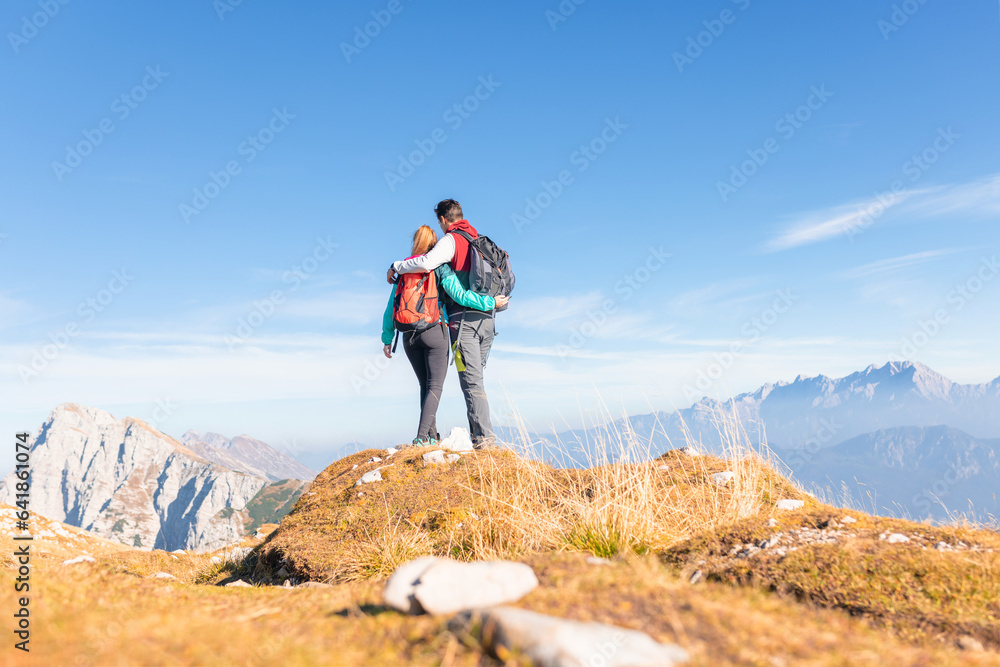 Young couple hikers hugging and looking at a fantastic mountain panorama, pointing in the distance on the bright sunny day Young couple hikers hugging and looking at a fantastic mountain panorama, pointing in the distance on the bright sunny day