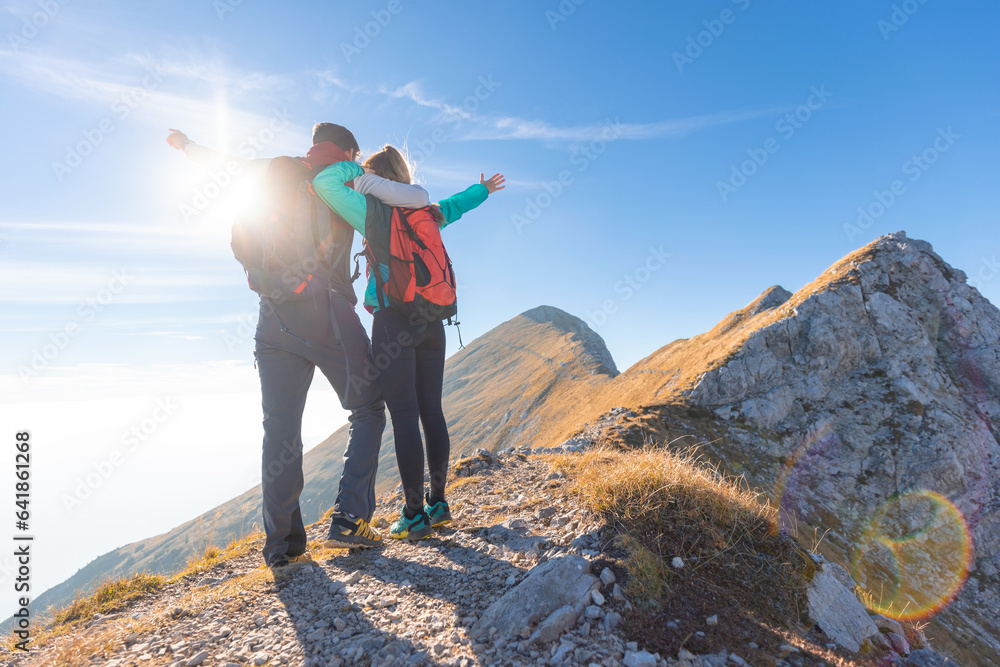Fantastic view of a hiker couple standing on a ridge celebrating success with spread arms, overlooking a magnificent mountain range landscape, aerial shot. Winner concept.