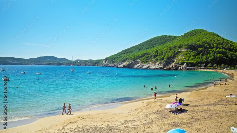 People having good time getting tan under sun at Playa De Santa Eulalia Beach. Tourists walking on the sand and relaxing at a beautiful beach in Ibiza, Balearic Islands, Spain. People having good time getting tan under sun at Playa De Santa Eulalia Beach. Tourists walking on the sand and relaxing at a beautiful beach in Ibiza, Balearic Islands, Spain.