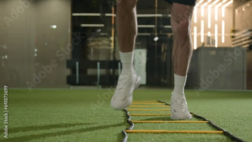 Unknown professional footballer running exercise jumping between ladder sticks male sportsman man soccer player muscular legs in sneakers fast jogging in sport gym training workout on football pitch