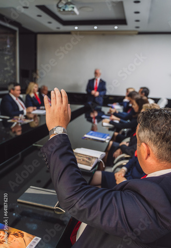 Businessman raising hand to ask question his elderly trainer during meeting in modern conference room.