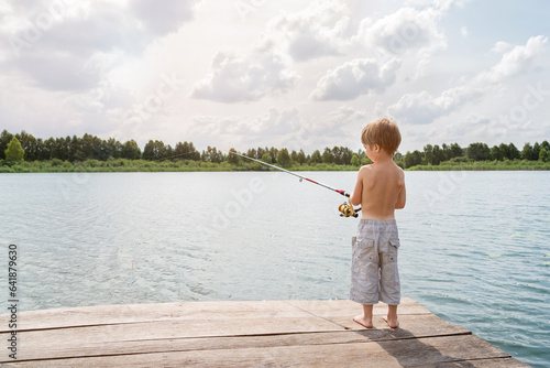 Boy fisherman catches fish in the lake. The child is standing on a wooden pier. The other shore of the lake is on the horizon. Sunny. Shooting against the sun.