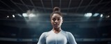 African American female gymnast in a stationary pose looking away from the camera with a faint sports arena in the background.