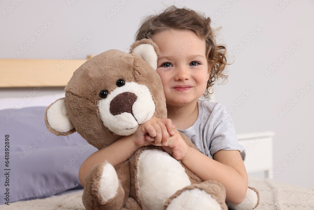 Cute little girl with teddy bear on bed indoors