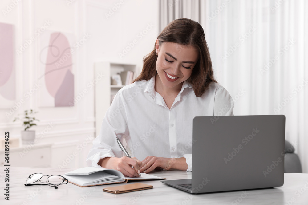 Happy woman writing something in notebook near laptop at white table in room Happy woman writing something in notebook near laptop at white table in room