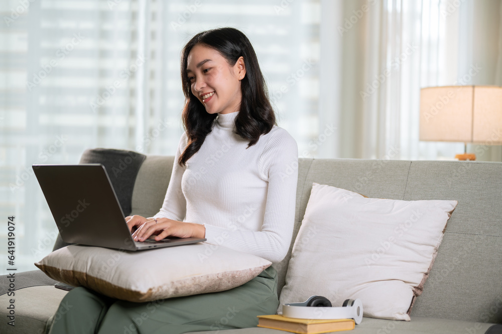 A beautiful Asian female is using her laptop on a sofa in her living room. Work from home A beautiful Asian female is using her laptop on a sofa in her living room. Work from home