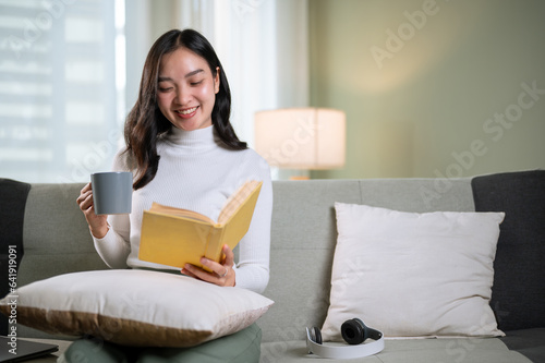 A beautiful Asian woman enjoys reading a book while having her morning coffee on a sofa