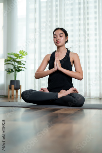 A beautiful and calm Asian woman in sportswear is meditating on a yoga mat in her living room