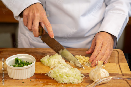 mãos de uma cozinheira usando Dolmã, preparando e picando alho e cebola sobre uma tabua de madeira e mais pote branco com salsa e cebolinha picados. photo