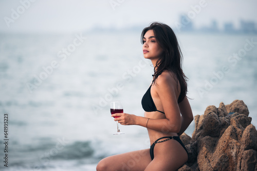 Portrait of young adult female Caucasian wearing black bikini and holding a glass of wine sitting on the rock seaside in vacation,