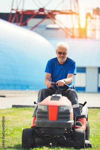 Worker mows territory of industrial facility. Man rides self-propelled lawn mower on lawn on summer sunny day. Elderly pensioner moonlights as gardener.