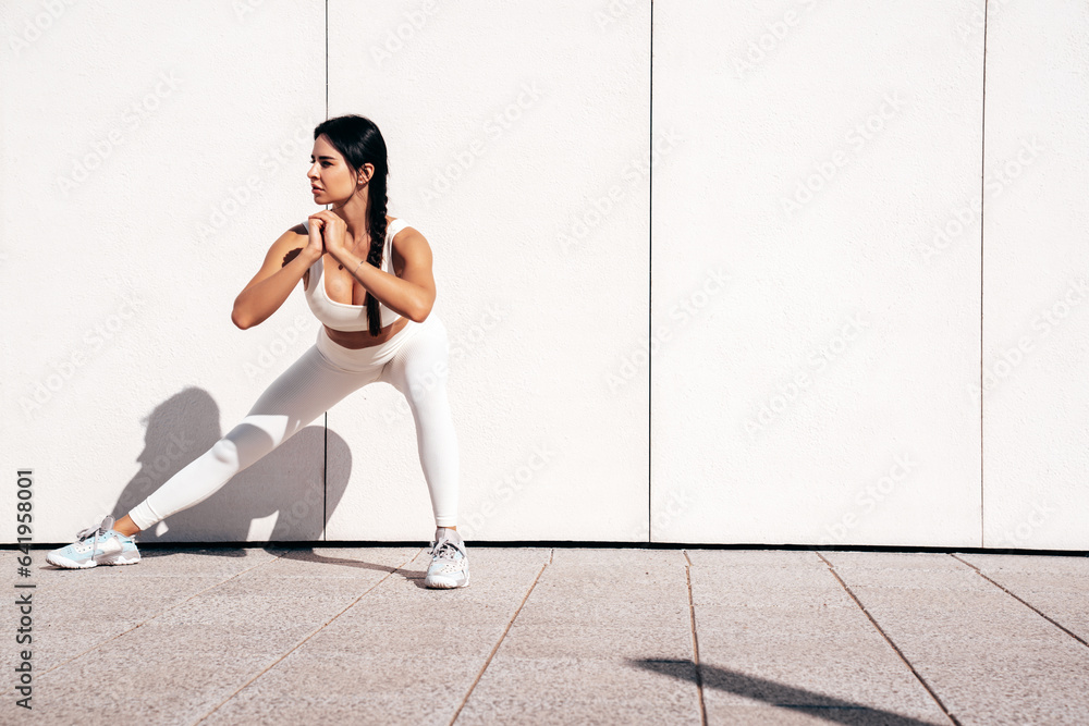 Fitness woman in sports clothing. Sexy young beautiful model athlete doing fitness workout. Female making exercises in the street at sunny day. Stretching out before training. Makes lunges near wall Fitness woman in sports clothing. Sexy young beautiful model athlete doing fitness workout. Female making exercises in the street at sunny day. Stretching out before training. Makes lunges near wall