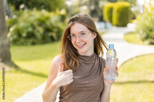 French girl with glasses holding a bottle of water at outdoors with thumbs up because something good has happened