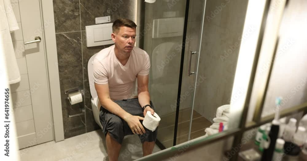 Bored man poops sitting on toilet bowl with toilet paper in hands in front of mirror in bathroom. Concept of natural process slow motion Bored man poops sitting on toilet bowl with toilet paper in hands in front of mirror in bathroom. Concept of natural process slow motion