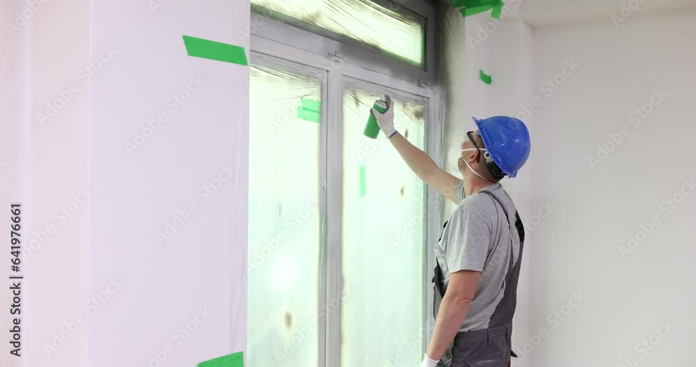 Helmeted builder spraying grey paint on window frame of apartment building. Handyman spraying grey paint. Interior renovation concept slow motion Helmeted builder spraying grey paint on window frame of apartment building. Handyman spraying grey paint. Interior renovation concept slow motion