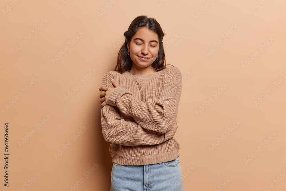 Pleased brunette Iranian woman daydreams embraces herself with both hands recalls romantic event keeps eyes closed smiles gently wears knitted jumper and jeans isolated over brown background. - obrazy, fototapety, plakaty 