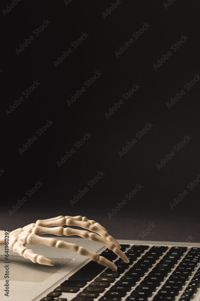 Fototapeta premium Vertical image of plastic skeleton hand on laptop keyboard with copy space on black background