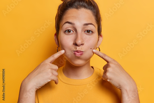 Fotografija Careless young woman with hair bun points index fingers at cheeks holds breath looks at camera dressed in casual t shirt isolated over vivid yellow background tries to be funny