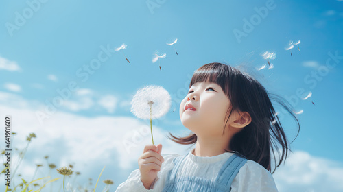 Photo of a little Asian girl blowing a dandelion, toddler child holding a dandelion flower on blue sky background, looking upwards, close-up summer portrait with copy space