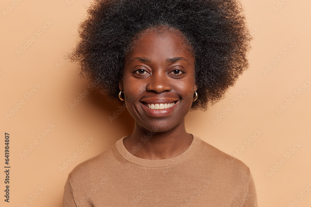 Portrait of beautiful dark skinned woman smiles toothily has curly bushy hair wears golden earrings and casual jumper isolated over brown background. People positive emotions and feelings concept Portrait of beautiful dark skinned woman smiles toothily has curly bushy hair wears golden earrings and casual jumper isolated over brown background. People positive emotions and feelings concept