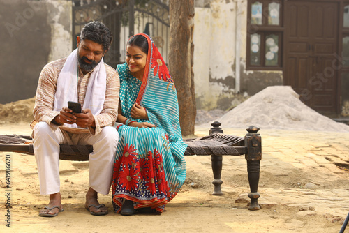 Happy Indian Rural family in village. couple sitting together outside their home front yard using mobile phone. Wife beautiful saree traditional wear